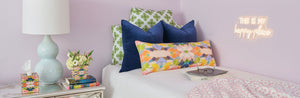 Decorative scene with pillows, lamp, and books on a bed against a purple wall.